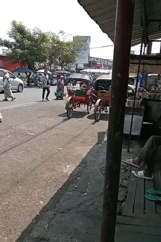 The video shows a street scene with a road and a sidewalk. On the road, several vehicles are visible, including a silver car, a black car, a motorcycle, and two pedicabs. One pedicab is red and green, and the other is brown. A person is walking on the sidewalk, and another person is sitting on a bench under a covered structure. The covered structure appears to be a market stall or a shop. There are trees and buildings in the background. The weather appears to be sunny.