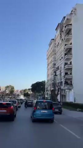 The video shows a street in what appears to be a city. There is traffic congestion with several cars lined up, with a blue van visible in the center of the frame. A motorcycle is navigating between the cars. On either side of the street are tall buildings, with one prominent white building to the right. In the distance is a park-like area with a large fountain spraying water. The sky is clear and blue. The lighting indicates it is daytime.
