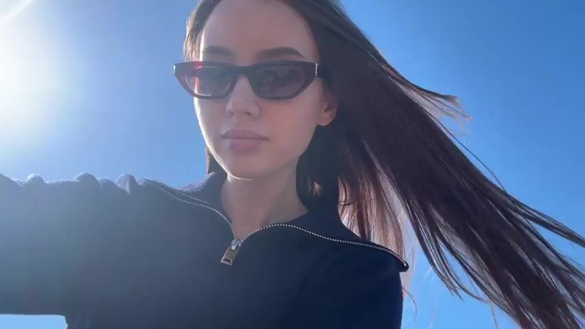 A woman is taking a selfie near the ocean on a sunny day. She is wearing dark sunglasses and a navy blue zip-up sweater. Her long brown hair is blowing in the wind. Behind her is a beach, a body of water, and a blue sky.