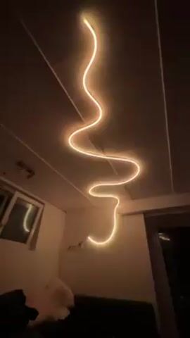 The video shows a light fixture on the ceiling changing colors. It begins as a soft orange with green accents, then transitions to full green, then a combination of green and blue, and finally to a solid blue. The light fixture is a long, curved tube that runs across the ceiling in a wavy pattern. In the background, there is a window and a partial view of a room, including a white pillow and a dark sofa. The walls are white.