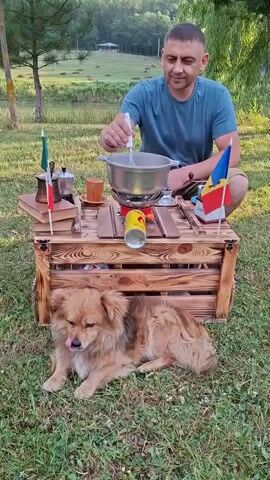 A man in a blue t-shirt is sitting cross-legged outdoors, stirring something in a pot on a portable stove. Beside him, a small, fluffy tan dog lies on the grass. A wooden crate serves as a table, holding the stove, the pot, a coffee maker, two cups, two books, a yellow can, and two small flags - one Italian and one Romanian. The man appears focused on his task. The setting is a grassy area with trees in the background and a body of water. The lighting suggests it is daytime, likely late afternoon or early evening, with soft sunlight filtering through the trees. The overall atmosphere is relaxed and domestic, perhaps a picnic or a casual outdoor cooking setup. The visible text on the yellow can is "Cola," a brand of soft drink. The scene is in Malcoci, Moldova.