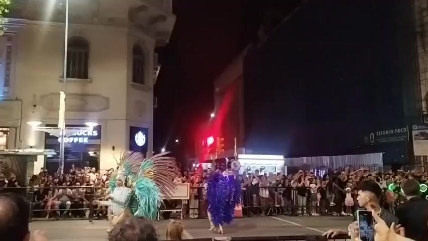 At night, the streets are crowded with people on both sides watching a parade. Several dancers in flamboyant carnival costumes, adorned with brightly colored feathers, are moving along the street. The crowd is mostly adults, and many are holding up their phones to record the event. A Starbucks Coffee shop is visible in the background. Streetlights illuminate the scene. There are barriers lining the street, keeping the crowd back from the performers.