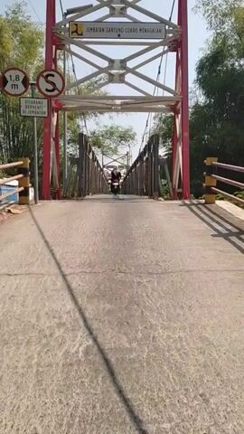 The video shows a first-person perspective of someone riding a motorcycle across a suspension bridge. The bridge is made of metal and has wire mesh railings. There are trees and foliage on either side of the bridge. In the distance, another motorcycle can be seen on the bridge. The sky is clear and sunny. A sign on the bridge reads "JEMBATAN GANTUNG COKRO MENGGALAN". There is also a circular traffic sign with a red border and a black "S" inside it. The camera is mounted on the front of the motorcycle and is angled slightly downwards, providing a clear view of the bridge and the road ahead.