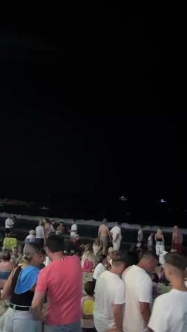 A wide shot captures a crowded beach at night. The sky is dark, with some distant lights visible over the water. A large group of people are gathered on the sand, some are standing, some are sitting in chairs, and some appear to be in the shallow water. Many people are dressed in light-colored clothing. The overall atmosphere appears to be social and relaxed. The video pans slightly to the right, revealing more of the beach and the people on it.