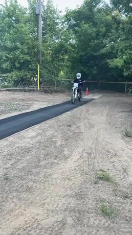 A person on a white dirt bike with a white helmet rides over a black ramp with red trim. They are on a dirt track in front of trees and a blue house. The person then rides up the ramp and jumps into the air over an inflatable landing pad. The person on the dirt bike is wearing a black shirt and pants and white helmet. The sky is clear and blue.
