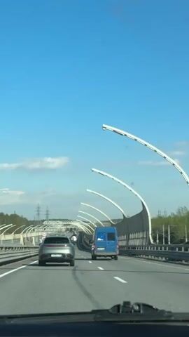 The video shows a drive on a highway during daylight. The weather is clear with a blue sky and a few clouds. The view is from the driver's perspective. Two vehicles are ahead: a silver car and a blue van. The highway has three lanes in each direction, divided by concrete barriers. There are distinctive white, arched structures along the sides of the highway.  Speed limit signs indicating 90 are visible. Utility poles and trees are visible in the distance. There is music playing in the background.