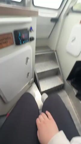 The video shows the interior of a moving boat. It starts with a view of the front portion of the cabin, including metal steps leading up to the driver's seat. The camera pans upward to show the boat's control panel and steering wheel, where a person is operating the controls. Then, the camera moves to show a woman sitting in a seat in the main cabin, looking at her phone. Other people can be seen sitting in the seats behind her. The overall environment appears to be clean and well-maintained. Outside the boat's windows, there is a view of a body of water and land in the distance.