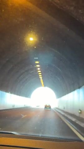 The video shows a car driving through a tunnel on a sunny day. The tunnel is lit by a row of lights along the top. As the car exits the tunnel, the road is surrounded by a railing and streetlights. The sky is blue with scattered clouds.