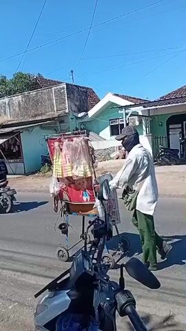 A person wearing a light-colored shirt, dark pants, and a face mask is pushing a stroller on a paved road. The stroller contains a doll or stuffed animal. The person is walking away from the camera towards the right side of the frame. The environment appears to be a rural or semi-urban area with buildings and trees visible in the background. The sky is clear and blue. A motorcycle is parked in the foreground, partially obscuring the view of the road.