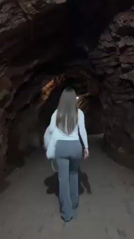 A woman is walking away from the camera down a dark tunnel. She has long brown hair and is wearing a white sweater and grey pants. She is carrying a white bag over her shoulder. The tunnel is lined with rock walls and has lights overhead. The floor of the tunnel is dirt.