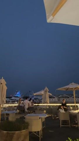 The video shows an outdoor dining area at dusk or night. The sky is a deep blue, and there are lights visible from boats in the background. The dining area has white umbrellas, tables, and chairs. Several people are seated at tables or standing and talking. In the foreground, there is a planter with greenery. In the background, multiple boats, some with lights, are docked at a marina. The lighting suggests a relaxed evening atmosphere.