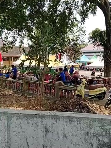 A group of people are gathered outdoors, some sitting and some lying down on a low wall or ledge. They are mostly dressed in casual clothing, with a few individuals wearing blue shirts. In the background, there are several colorful tents, suggesting an event or market. A yellow scooter is parked on the right side of the frame. The environment appears to be a park or open space with trees providing shade. The overall atmosphere seems relaxed, with people resting or observing.