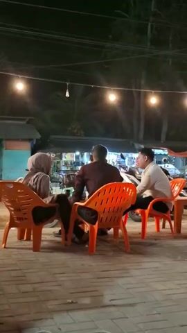 The video shows a nighttime scene at an outdoor cafe or eatery, likely in Keudeteupinpunti, Indonesia. Three people are seated at orange plastic chairs around a table. One person, wearing a hijab, faces away from the camera. Another person, wearing a dark shirt, also faces away. A third person, in a light-colored shirt, is seated to the right, facing the other two. In the background, several motorcycles are parked, and a small food stall or van is visible with the text "Naban Coffe" and "Samudera" printed on its side. Strings of lights are strung overhead, illuminating the area. The ground is paved with brick-like tiles. A truck passes by in the distance, its lights flashing. The overall atmosphere appears relaxed, with people gathering to socialize in the evening.