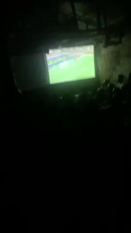 A group of people are gathered in a dark room, silhouetted against the bright glow of a projected image. The projection appears to be a sporting event, likely a soccer match, with players on a green field and visible advertisements on the screen. The room has a rustic, unfinished appearance, with exposed beams and rough walls. One person in the foreground is standing and facing the screen, their silhouette clearly defined. The overall atmosphere is one of shared viewing and engagement with the projected content.