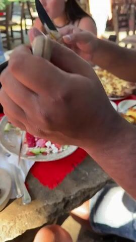 The video shows a close-up view of hands slicing a long, grilled sausage on a wooden board. The sausage appears to be well-cooked, with visible grill marks. The knife is sharp and used with precision, creating evenly spaced slices. In the background, a table is set for a meal, with plates of food, drinks, and other utensils visible. One plate contains what appears to be grilled eggplant or zucchini, and another holds a salad. People are blurred in the background, suggesting a social gathering or meal. The lighting indicates an outdoor setting, possibly a picnic or barbecue, with natural light filtering through trees. The focus is on the act of preparing the food, highlighting the texture and juiciness of the sausage as it is sliced. The overall impression is one of a relaxed, outdoor meal shared among friends or family.