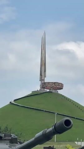 A static, medium shot captures a tall, triangular monument situated atop a green, grassy hill. The monument, constructed from concrete and metal, features three vertical spires and a circular band adorned with relief sculptures of human faces. A winding staircase ascends the hill towards the monument, with several small figures of people visible near the top. In the foreground, the barrel of a dark green tank extends into the frame from the bottom left, its muzzle facing right. The sky above is a pale blue with scattered white clouds. The overall scene appears to be an outdoor memorial or monument site, possibly with a military theme given the presence of the tank.