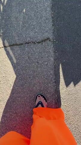 The video shows a first-person perspective of someone walking on a paved road. The person is wearing orange pants and black flip-flops. The sun is casting shadows on the road, indicating it is daytime. In the distance, there are houses, trees, and a few other people walking. A black car is also visible further down the road. The environment appears to be a residential street in a sunny climate.