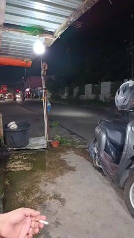 The video shows a nighttime street scene from a covered outdoor area. A person's hand holds a lit cigarette in the foreground, to the left. To the right, a parked scooter with a helmet on its seat is visible. A large black bin is next to a metal pole supporting the covered area. A small potted plant sits near the pole. Beyond the covered area, a street with traffic is visible. Headlights and taillights of moving vehicles are illuminated. A person on a bicycle is blurred as they pass by in the middle ground. The ground appears wet and reflective. The overall environment suggests a street-side establishment or resting point.