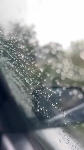 The video captures a close-up view from inside a car, looking out through a windshield covered in raindrops. The water droplets are prominently featured, creating a bokeh effect against the blurred background. A portion of the car's side mirror is visible in the lower right corner of the frame. The indistinct background shows what appears to be trees or foliage and a road, indicating an outdoor setting during rain. The weather is clearly overcast and rainy. Upbeat music with foreign language vocals plays in the background. No text is visible in the video. The location context provided is Marino Pole, Bulgaria.
