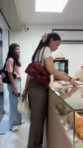 The video shows the interior of a bakery. A woman wearing a white shirt and brown pants stands at a counter, seemingly placing an order with a bakery employee wearing a tan apron and white t-shirt. Another woman in a pink top and jeans stands to the side, holding a white bag. A display case in front of the counter holds various pastries, including croissants and almond croissants. Behind the counter, another bakery employee, a young man with a similar apron, is preparing something, likely coffee, on a counter with various coffee-making equipment. The bakery has white walls and bright lighting. There is an air conditioning unit on the wall above the counter. The camera moves slightly to the right to capture the scene.