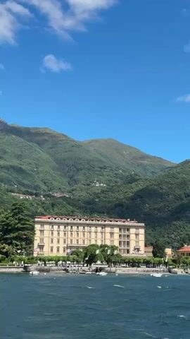 This vertical video captures a picturesque lakeside scene in Loveno, Italy, under a clear, sunny sky. The perspective appears to be from a moving boat on the lake, indicated by the slightly choppy water in the foreground and the gentle camera movement.

In the midground, a grand, multi-story building with a light beige or cream-colored facade and a red-tiled roof stands prominently along the waterfront. This structure, possibly a hotel or large residence, features multiple windows and balconies across its various levels. Several Italian flags, displaying green, white, and red vertical stripes, are visible hanging from the building or along the promenade in front of it. A tree-lined walkway separates the building from the water's edge, where a few people can be seen strolling. Mature green trees, including a large evergreen on the left, frame the scene.

Numerous small, white-hulled boats are moored or anchored in the dark blue-green lake water closer to the shore.

The background is dominated by majestic, verdant mountains, covered in dense forestation. These steep slopes rise dramatically under a bright blue sky dotted with scattered white clouds, creating a stunning natural backdrop for the lakeside architecture. The overall impression is one of serene beauty on a pleasant, sunny day.
