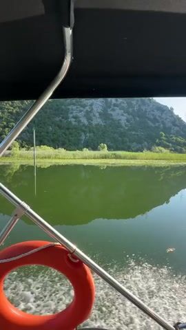 The video shows a boat moving through calm water. A life preserver is visible in the foreground, attached to a railing. The boat has a dark canopy overhead. A small blue flag with white lettering, possibly "SIDE," is attached to the canopy. The boat is leaving a white wake behind it. The surrounding environment is a body of water reflecting green foliage and hilly, mountainous terrain under a clear blue sky. The camera appears to be mounted on the boat, looking backward towards the water and the landscape. The engine of the boat, a black Suzuki outboard motor, is partially visible in the frame. The water is a muted green color, possibly due to algae or vegetation. The hills are covered in lush green trees and shrubs, with rocky outcrops visible. The sky is a vibrant, cloudless blue. The overall impression is one of peaceful boating on a scenic lake or river.