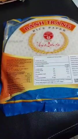 The video shows a close-up of a package of Banh Trang Rice Paper. The package is blue with orange and white text and images. The text is in Vietnamese and English, and it includes nutritional information and ingredients. The rice paper is round and white. The package also features an image of fresh spring rolls and a bowl of sauce. A hand is seen briefly touching the package in the first few seconds of the video. The video appears to be filmed in an indoor setting with a dark background.