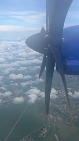 From the perspective of an airplane window, a rotating black propeller with yellow tips, part of a blue engine, occupies the right side of the frame. The plane is flying over a landscape with green fields, patches of white, fluffy clouds, and visible infrastructure like roads and buildings. The sky above is light blue with scattered clouds. The ground below shows a mix of rural and possibly semi-urban areas, with a highway visible stretching across the land. The scene depicts a daytime flight with good visibility, offering an aerial view of the terrain.