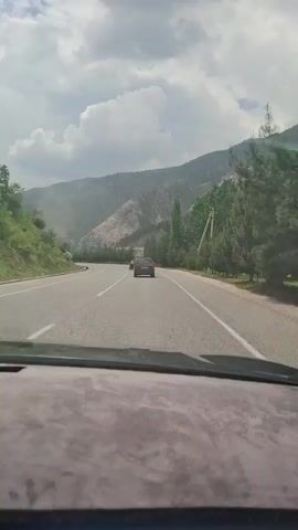 The video shows a car driving on a two-lane road in a mountainous area.  The road curves to the left. There are trees lining the side of the road on the right. Mountains are visible in the distance, with some areas appearing to have been excavated.  Another car is driving ahead on the same lane. The sky is cloudy.
