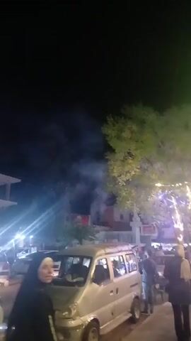 The video shows a nighttime scene with fireworks being set off. There are people gathered in the street, some standing near cars. There is a tree with lights on it. Smoke from the fireworks drifts across the scene. The camera moves slightly, following the fireworks.
