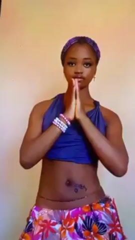 A young woman with dark skin stands facing the camera, wearing a blue halter top, a brightly colored floral skirt, a beaded headband, multiple bracelets, and a beaded waist chain. She has her hands pressed together in front of her in a prayer position, her gaze level and direct. The background is a plain, light-colored wall. The lighting is consistent, allowing clear visibility of the subject. She appears to be centered in the frame, with no significant distractions around her.