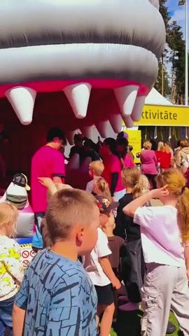 A group of adults and children are gathered around the entrance of a large inflatable bouncy house designed to look like a giant crocodile or shark's mouth with large pink and white teeth. Some people are entering and exiting the bouncy house, while others are observing from the outside. The scene takes place outdoors on a sunny day, with green grass and trees visible in the background. A yellow sign with the text "AKTIVITĀTE" is visible behind the bouncy house. Some people are wearing casual clothes suitable for summer weather.