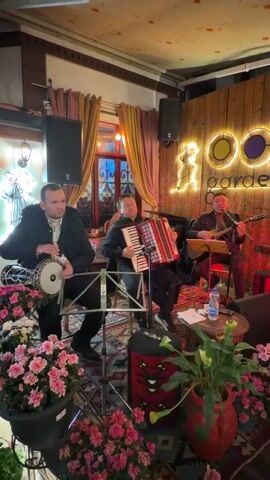 Three men are playing musical instruments in what appears to be a bar or lounge. The man on the left is playing a doumbek, the man in the middle is playing an accordion, and the man on the right is playing a guitar and singing. They are all sitting on chairs, and music stands with sheet music are in front of the accordion player and guitarist. In the foreground, there are several pots of pink flowers. There is also a patterned rug under the band. Behind the band, there is a wooden wall with a lighted sign that says "OO garde." There are also speakers and a window with red and orange curtains.