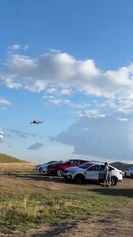 A drone flies in the sky above a parking lot filled with cars and people. The drone is in the center of the frame, and it is flying at a moderate speed. The drone is a dark color and has four propellers. The drone is in focus, and the background is slightly blurred. The background is a blue sky with white clouds. There are some trees and mountains in the distance. There are several cars parked in the parking lot. The cars are all different colors and models. There are a few people standing around the cars. The people are talking and looking at the drone. The drone is a DJI Mavic Mini or similar model.