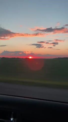 The video shows a sunset over a field. The sky is a mix of blue, orange, and pink, with some clouds scattered across it. The sun is a bright red orb just above the horizon. A strip of dark gray road is visible in the foreground, blurred from motion. The field appears to be green and vast, stretching to the horizon. The overall impression is that the video was filmed from a moving vehicle.