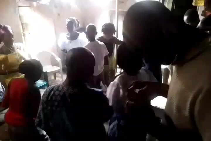The video shows a group of people gathered indoors, possibly in a home setting. There are at least eight individuals visible, including women and children. One woman is wearing a yellow dress with an elaborate purple headwrap, while others are wearing white tops. Some individuals appear to be standing, while others are seated. A patterned cloth is draped over a surface in the foreground.  The room is lit by natural light coming through a window. It is difficult to discern specific actions or interactions among the group due to the video's perspective and the movement of the camera.
