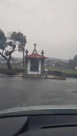 The video shows a roadside shrine under a gray sky, filmed from inside a car. The shrine is small, square, and white, with a red-tiled roof and a cross on top. There are decorative finials at each corner of the roof. The shrine appears to house a statue or image behind glass. The road in front of the shrine is wet, likely due to rain. To the left of the shrine is a tree, and in the background is a view of a town or village, with various buildings visible. The overall atmosphere is somber and overcast. The camera is stationary inside the car.