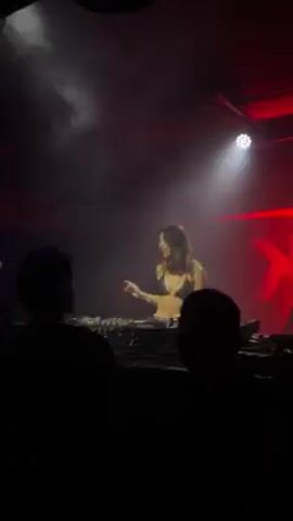A female DJ wearing a bra and long hair is performing in a dimly lit club with a fog machine. A red neon light is visible on the back wall. Two people are in the foreground facing the DJ, seemingly watching her performance. She is working on her equipment.