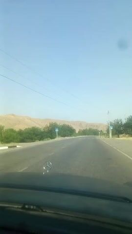 A video begins with a view from inside a vehicle, looking out through the windshield at a two-lane asphalt road stretching into the distance. The road is flanked by sparse vegetation and dry, hilly terrain under a clear blue sky. The hood of the car, with a Mercedes-Benz emblem, is visible in the foreground. On the side of the road, two pedestrian crossing signs with blue squares and white figures are visible at intervals. The vehicle appears to be moving at a moderate speed, indicating travel. The environment suggests a rural or semi-arid setting. The video is short, lasting only a few seconds and showing a continuous, unchanging scene of the road ahead.