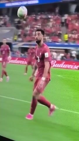 This video shows a soccer match. The focus is on a player in a maroon jersey with the number 8 on his back, who is heading a soccer ball. The player is on a green field, with spectators in the background. The action appears to be taking place during a game, as other players in similar jerseys are visible on the field. The camera is positioned to capture the player's action from a slightly elevated angle. The lighting suggests it is daytime and possibly indoors or under stadium lights. The image quality is somewhat pixelated.