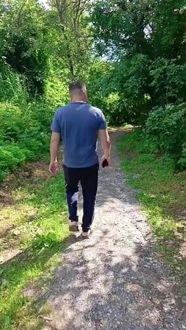 A man walks away from the camera down a gravel path through a wooded area. He is wearing a dark blue t-shirt and dark pants with a white stripe down the side. There is green foliage on either side of the path. The sun is shining through the trees, casting shadows on the path.