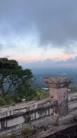 From an elevated perspective, a concrete balcony overlooking a green and hilly landscape can be seen. The sky is overcast with hues of pink near the horizon. Vegetation grows from the concrete structure of the balcony, including grass and small trees. The distant landscape shows signs of habitation, with glimpses of buildings and smoke plumes. The camera slowly pans to the right, revealing more of the landscape and a tall tree silhouetted against the sky.