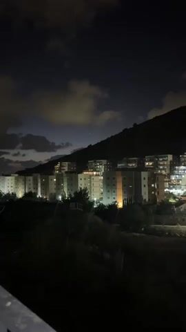 The video shows a nighttime view of a hillside covered in buildings. The buildings are apartment complexes with lights visible in many windows. Trees and foliage are present on the lower slopes of the hill. The sky is dark with some clouds illuminated by the lights below. There are no people or discernible actions visible in the video. The scene appears to be a residential area at night.