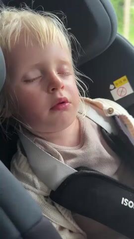A close-up shot shows a young child with blonde hair sleeping in a car seat. The child is wearing a light-colored shirt and a cream-colored cardigan. The car seat is black, and the child is secured with a harness. A yellow sticker with black symbols is visible on the car seat. The lighting suggests it is daytime.