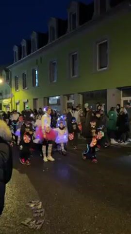 In a dim outdoor setting, possibly twilight, a group of people, including children and adults, are seen walking down a wet street. Many individuals are dressed in winter clothing like jackets and hats. A few children are dressed in colorful, festive outfits, including pink tutus and light-up accessories. Some participants carry illuminated objects, perhaps lanterns or decorations, adding to the ambiance.  The street is lined with buildings, one of which is a distinct green. The people appear to be part of a parade or celebratory procession.  A muffled booming sound is heard.  The camera appears to be held by someone walking with the group.