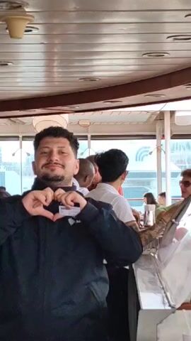 Here is a brief description of the video:

The video takes place on what appears to be a boat or ferry, possibly a tourist vessel, with a bar area. The primary focus is on a bar worker wearing a black jacket. He is facing the camera and makes a heart symbol with his hands. He is smiling and seems friendly.

The scene then shifts to the bar area with other bar service workers. One worker is wearing a white shirt with "BAR SERVICE" printed on the back. He has tattoos on his arms. Several customers are standing at the bar. Another bartender is wearing a white shirt and black pants. He is talking to a customer at the bar. There are bottles and glasses behind the bar. Outside the windows, a cityscape is visible.