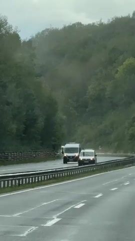 The video shows a highway with multiple lanes of traffic. It appears to be raining, as evidenced by the water droplets on the camera lens and the wet road surface. Several vehicles are visible, including white vans, a black car, and a red car. The vehicles are traveling in the same direction, with some of them in a convoy. Trees and foliage line the sides of the highway, suggesting a rural or wooded area. The overall impression is of a cloudy, possibly overcast day with limited visibility due to the rain. There are no discernible people in the video apart from blurry figures within the vehicles. The camera angle suggests the video was filmed from inside another moving vehicle.