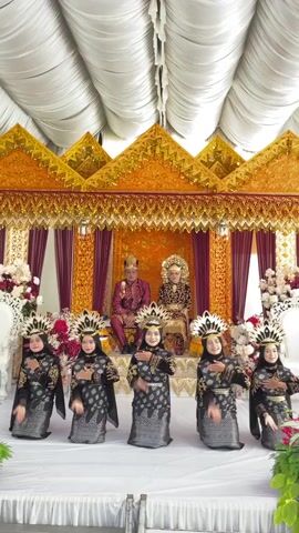 This is a video of a traditional wedding ceremony in Kulam Baro, Indonesia. The scene is set on a stage decorated with ornate golden structures and deep red curtains. A bridal couple is seated on a raised platform, dressed in elaborate traditional attire. The bride wears a golden headdress and gown, while the groom is in a maroon outfit.

In front of the couple, six young girls are performing a dance. They are all dressed in matching black and silver traditional outfits, complete with striking headdresses. They appear to be performing a synchronized dance routine with graceful hand movements.

The overall atmosphere is festive and celebratory, typical of a wedding event. The lighting suggests it's daytime, likely indoors or under a tented area given the draped white ceiling. The setting is richly decorated, emphasizing the cultural significance of the occasion. There is no visible text in the video.