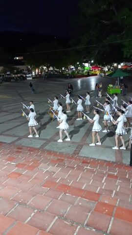 At night in an outdoor public space, a marching band is performing a show. The band is comprised mostly of young women, each dressed in a white skirt and top, and a sailor hat. They are carrying illuminated batons that are emitting a rainbow of light. At the head of the band is a man in a similar uniform. A woman in a patterned shirt and black pants is walking along with the band, presumably their instructor. The group is on a light-colored stone square that is decorated with a diamond pattern. A set of brick stairs leads up to the square. The square is surrounded by trees and other people. There are food vendors under umbrellas, and benches with people sitting on them. In the background is a large sign that reads “YO TIJANA”.