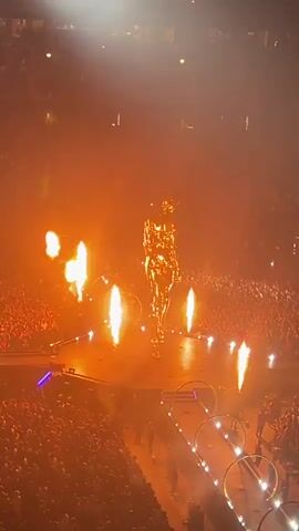 A person in a golden, metallic suit stands on a stage in a large arena. The suit appears to be a full-body costume, reflecting the stage lights. Around the performer, there are several pyrotechnic effects, including bursts of flame shooting upwards and outwards. The stage is elevated and appears to be part of a concert or performance setup, with circular metallic structures and lighting elements visible. The audience fills the seats of the arena, blurred in the background due to the lighting and distance. The overall atmosphere is dramatic and energetic, with a lot of orange and yellow light from the flames and stage lighting illuminating the scene. The camera angle is from a high vantage point, looking down at the stage.
